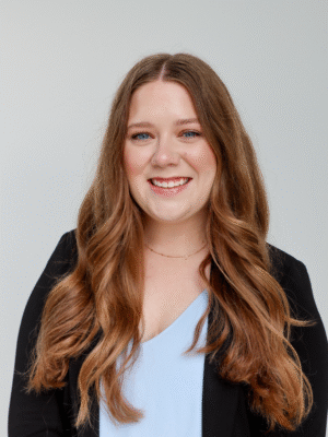 A young woman with long wavy brown hair, wearing a black blazer over a light blue top, smiles in front of a plain light background—perfectly dressed for Special Events.