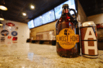A growler and a can with "Almost Home Taphouse" branding sit on a bar counter in Little Elm; menu screens and craft beer taps are visible in the background.