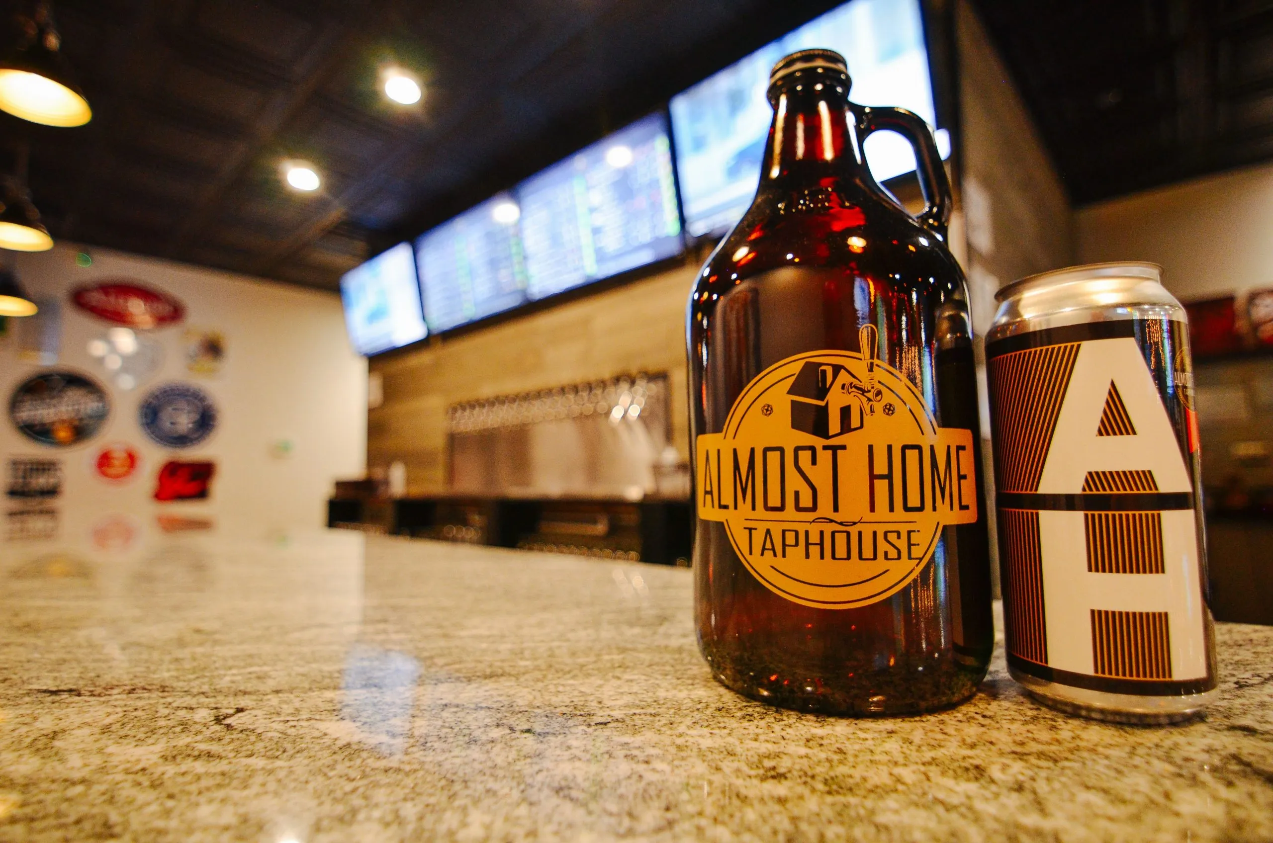 A growler and a can with "Almost Home Taphouse" branding sit on a bar counter in Little Elm; menu screens and craft beer taps are visible in the background.