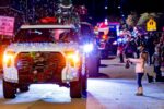 A girl stands on the sidewalk holding a bag as decorated trucks with Christmas lights and wreaths pass by in a nighttime parade.
