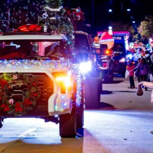 A girl stands on the sidewalk holding a bag as decorated trucks with Christmas lights and wreaths pass by in a nighttime parade.