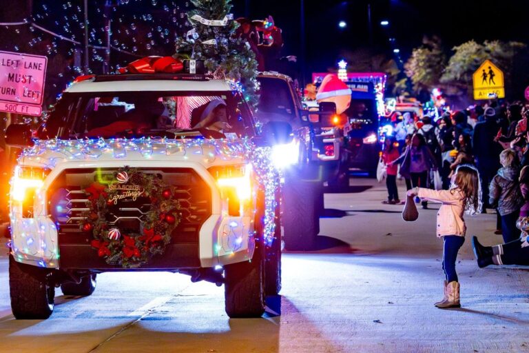 A girl stands on the sidewalk holding a bag as decorated trucks with Christmas lights and wreaths pass by in a nighttime parade.