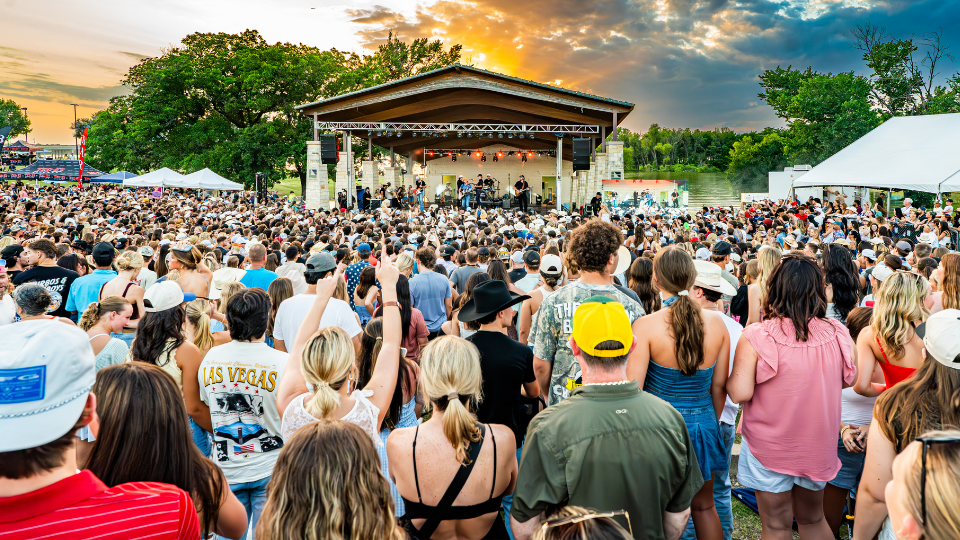 A large crowd gathers outdoors in front of the Brew & Que stage, enjoying a live band performing at sunset, surrounded by trees, tents, and the aroma of sizzling BBQ.