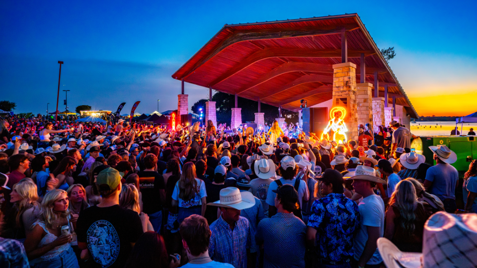 A large crowd in cowboy hats gathers at an outdoor stage during sunset, enjoying a live music performance under a wooden pavilion with plenty of bbq and cold beer at the Brew & Que festival.