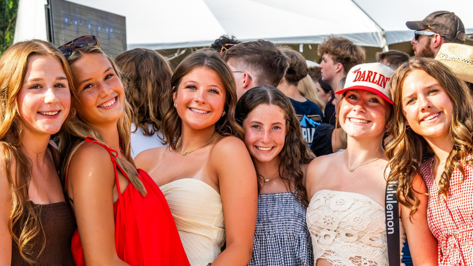 Six young women pose and smile for a photo at Brew & Que, an outdoor event with a lively crowd, beer in hand, and tents visible in the background.