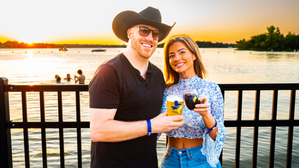 A smiling man in a cowboy hat and a woman hold drinks by a lakeside fence at sunset during Brew & Que, with people swimming in the background and the atmosphere filled with classic bbq vibes.