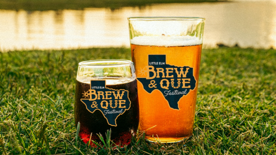 Two festival-branded Brew & Que glasses, one filled with beer and the other with a light drink, sit on grass near water at sunset.