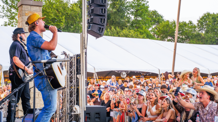 A musician with a guitar performs on an outdoor stage in front of a large, enthusiastic crowd near a white event tent at the lively Brew & Que, where fans enjoy great music, beer, and BBQ.