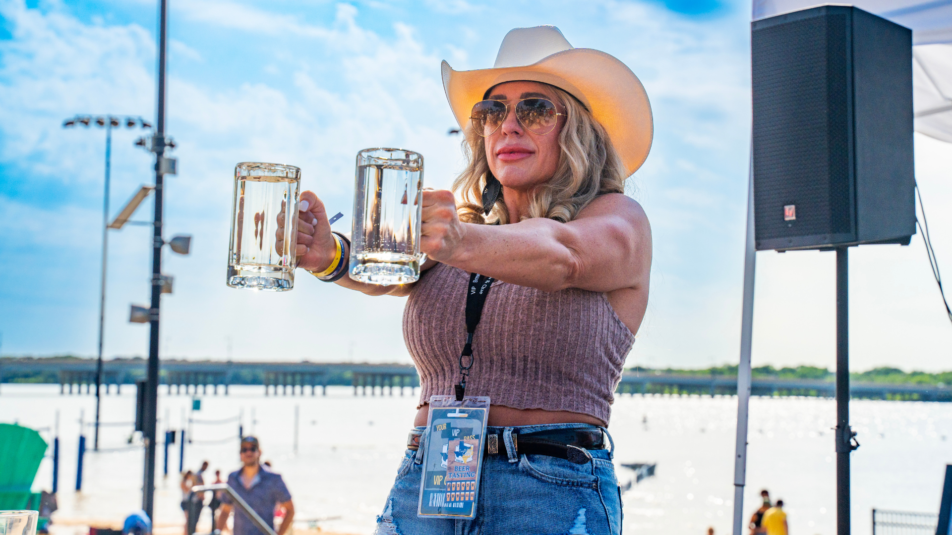 A woman in a cowboy hat and sunglasses holds two large glass mugs of beer with outstretched arms at an outdoor waterfront Brew & Que event.