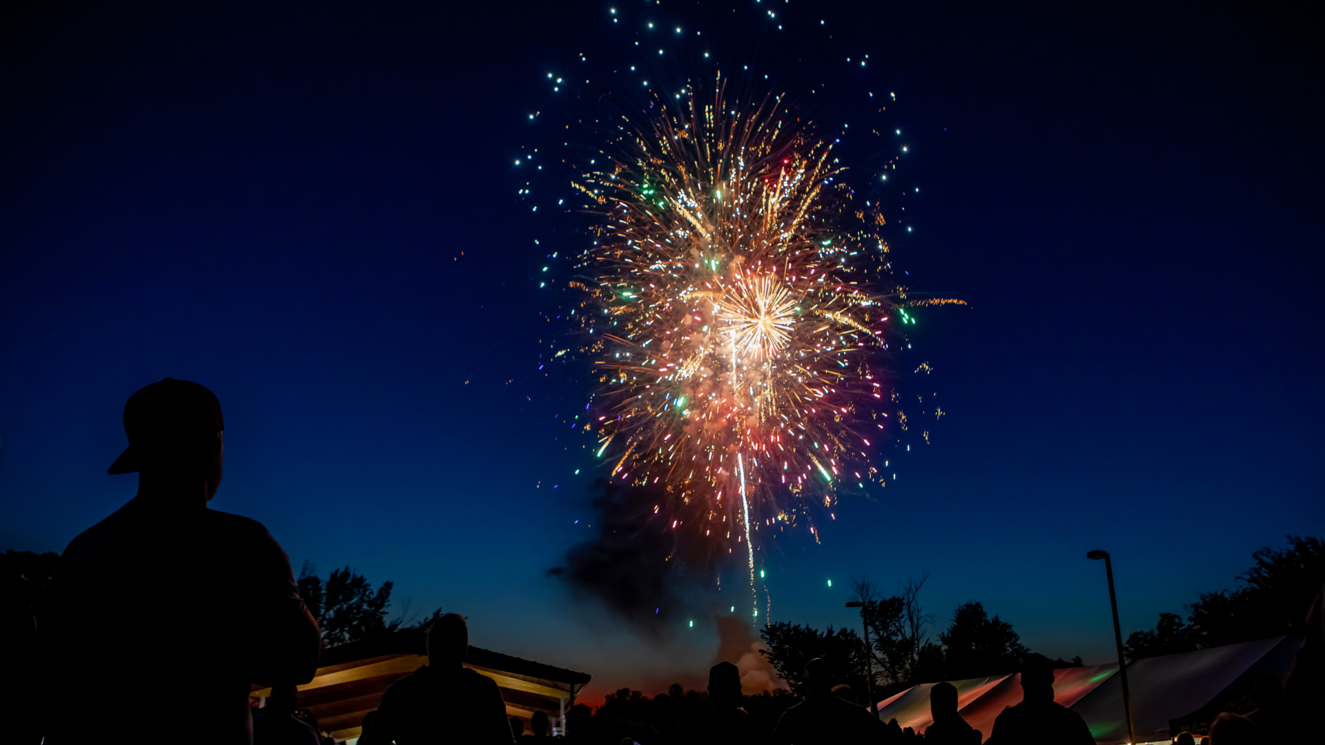 Colorful fireworks burst in the night sky above silhouetted people and buildings during an outdoor Brew & Que event, adding excitement to an evening filled with bbq and fun.