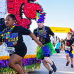 Children in colorful Mardi Gras attire participate in a festive race, sprinting past a cajun-decorated float and a yellow finish line arch on a sunny Big Easy day.