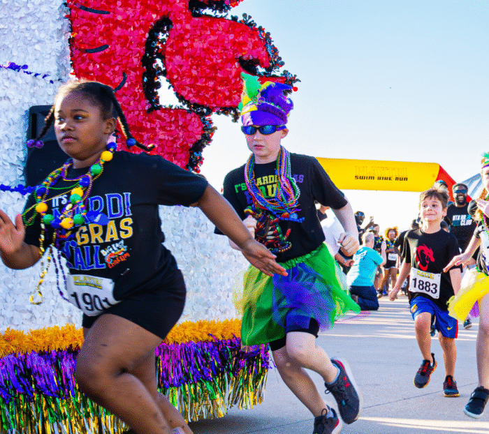 Race Children in colorful Mardi Gras attire participate in a festive race, sprinting past a cajun-decorated float and a yellow finish line arch on a sunny Big Easy day.