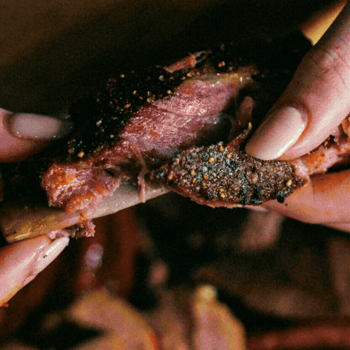 Close-up of hands pulling apart a seasoned, cooked piece of meat from a bone at Brew & Que BBQ—perfect for pairing with an ice-cold beer.