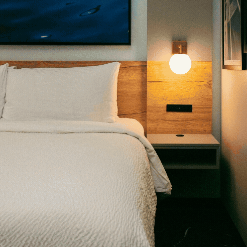 A neatly made bed with white linens next to a wooden headboard, a wall-mounted lamp, and a small nightstand—perfect for unwinding after a day at Brew & Que enjoying great beer and BBQ in this modern room.