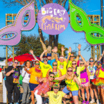 A group of people in colorful outfits pose under a "Big Easy in Little Elm" sign with large green and purple masks, bringing vibrant Cajun flair to this lively outdoor event.