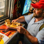 A man in a striped shirt and red cap picks up a tray of Cajun fries from a food truck window at night, savoring the bold flavors of the Big Easy.