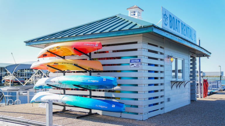 A boat rental shack with kayaks in various colors, including yellow, orange, red, and blue, displayed on racks outside. The shack has a sign reading "Boat Rental" and is located near a marina.