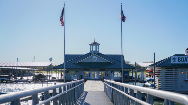 MARINA A waterfront building with a lighthouse-style cupola is visible at the end of a walkway. Two flags are flying on tall poles; the American flag and another flag. Boats and covered docks are seen on either side of the walkway under a clear blue sky.