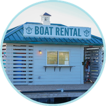 RENTAL (1) A small building with a sign reading "Boat Rental" on the roof. It has a window and entryway, and is situated by the water.
