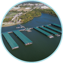 SLIP (1) Aerial view of a marina with multiple covered docks extending over a body of water. In the background, there is a sports complex with a baseball field and surrounding buildings. A residential area can be seen further in the distance.