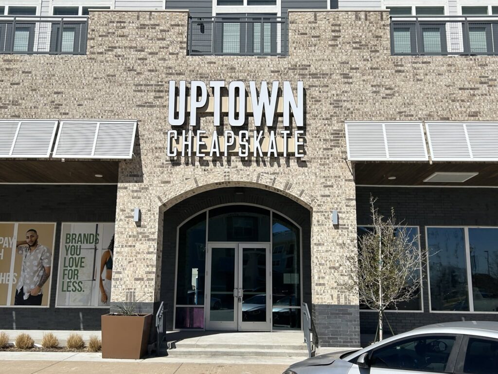 A storefront with a brick exterior features a large sign reading "Uptown Cheapskate." The entrance has glass doors beneath an archway. There is a poster to the left of the entrance displaying fashion models and text about brand value.