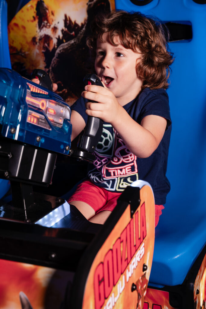 A child is enthusiastically playing a Godzilla-themed arcade game with two joysticks, seated in a gaming chair. The game has vibrant colors and Godzilla imagery.