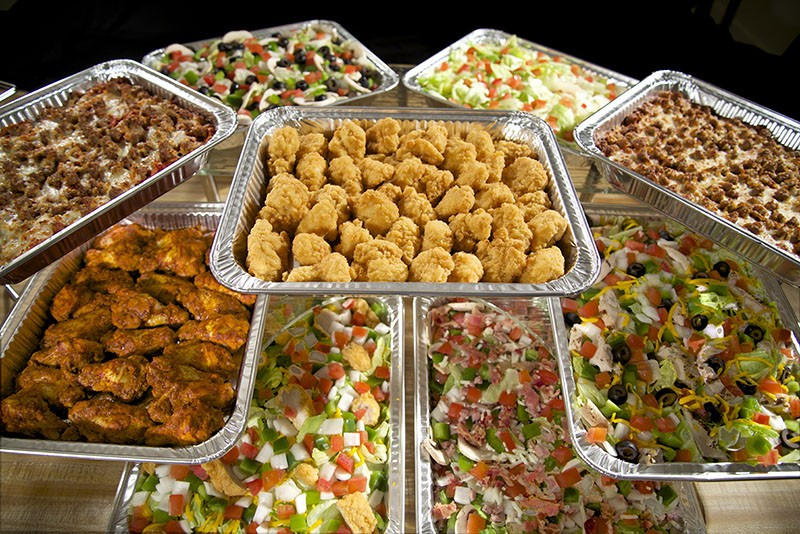 A variety of dishes in foil trays on a table. The dishes include fried chicken nuggets, saucy chicken wings, nachos topped with olives and tomatoes, a salad with cucumbers and cherry tomatoes, and a Mexican-style dish with diced vegetables and cheese.