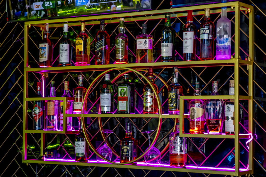 A well-organized bar shelf filled with various liquor bottles includes brands like Jack Daniel's, Jägermeister, and Ciroc, illuminated by pink and purple lights. A television screen above displays a sports game with scores from different teams.