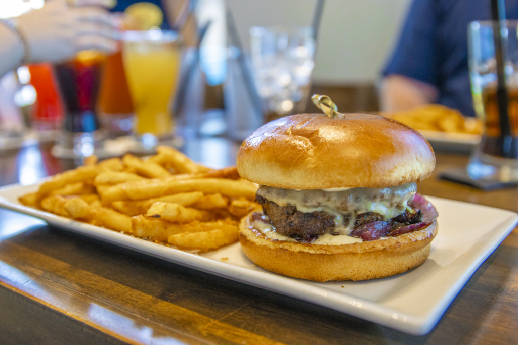 A close-up of a cheeseburger on a rectangular white plate, accompanied by seasoned fries. The background shows blurred drinks in various glasses and people seated at a wooden table.