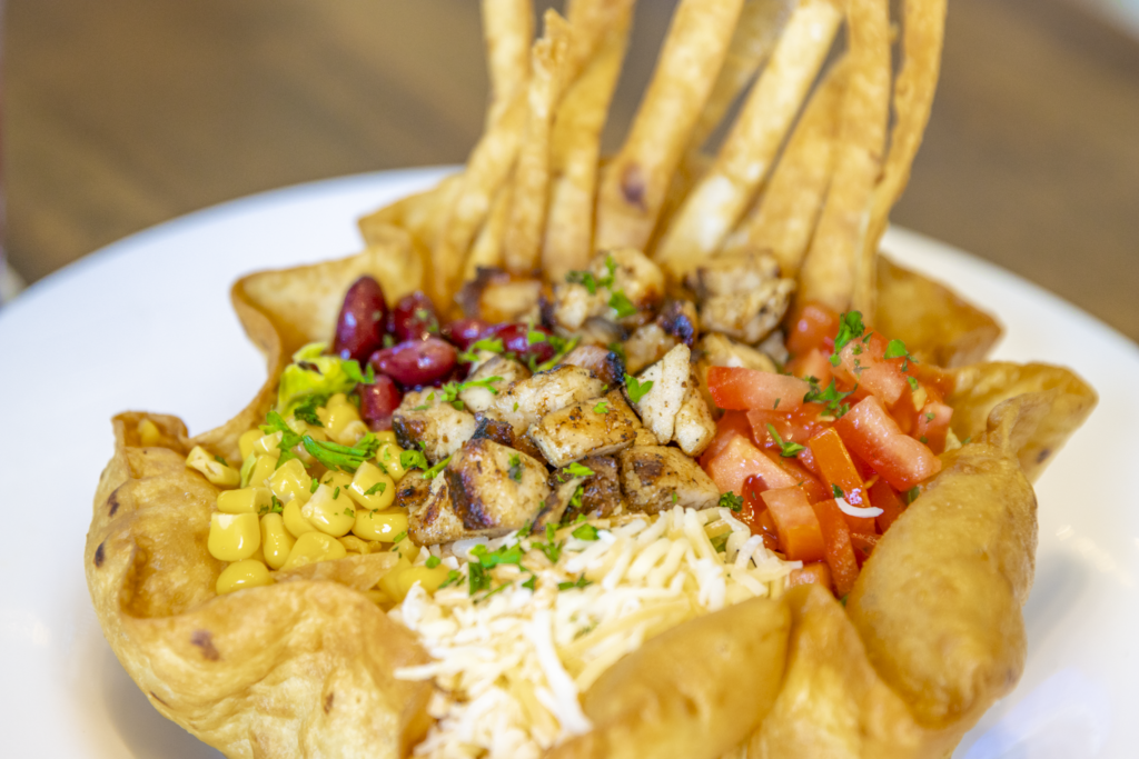 A vibrant salad is served in a crispy tortilla bowl on a white plate, containing grilled chicken, corn, kidney beans, diced tomatoes, and shredded cheese with thin tortilla strips.