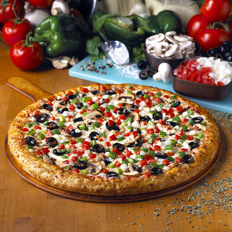 A freshly baked pizza topped with olives, mushrooms, green peppers, and diced tomatoes is on a wooden board. Surrounding the pizza are fresh vegetables like tomatoes, bell peppers, mushrooms, and onion, along with a small bowl of diced onions and tomatoes. The scene is set on a wooden table.