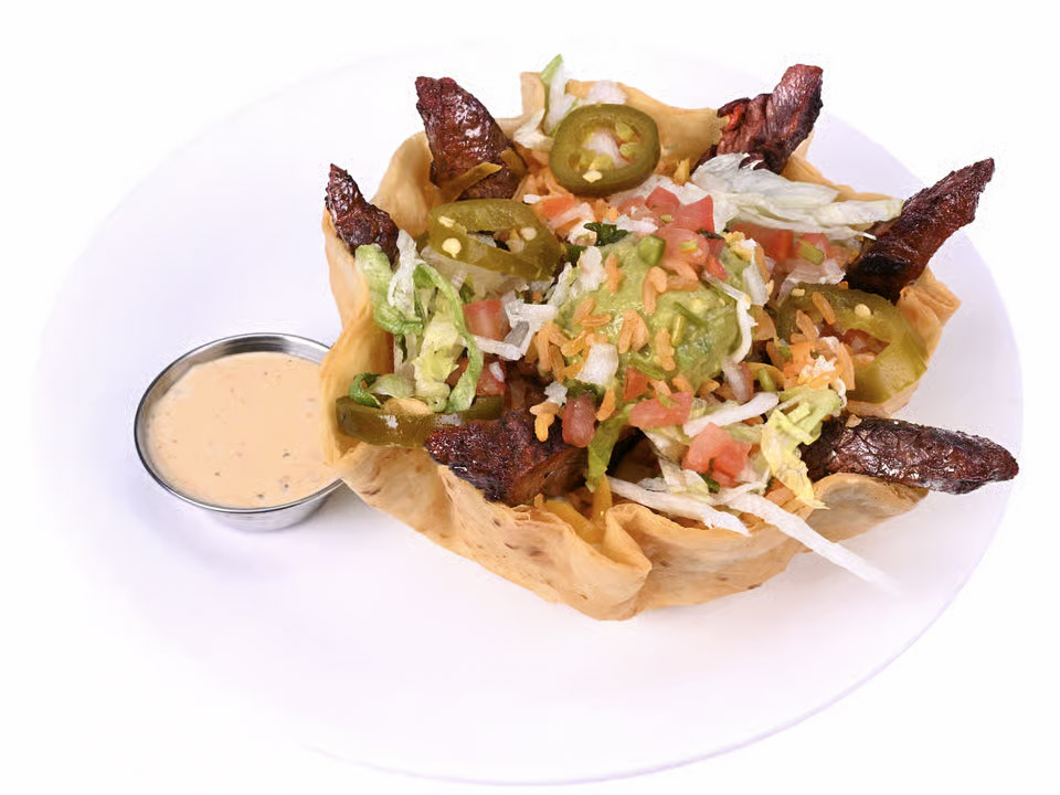 Fajita salad with steak, guacamole, jalapeños, tomatoes, and lettuce all in a friend corn tortilla bowl, with a side of creamy dressing.