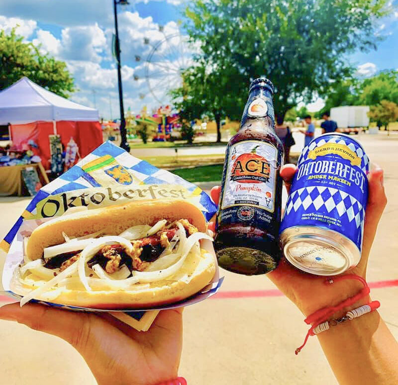Person holding an Oktoberfest sausage sandwich, an ACE Pumpkin beer bottle, and an Oktoberfest München can at an outdoor festival.