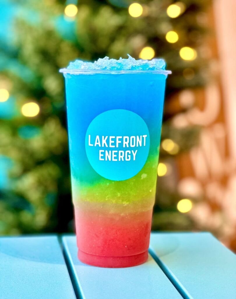 A tall plastic cup filled with a layered blue, green, and red drink sits on a light blue surface with a blurred festive background of glowing lights and greenery. A bright cyan sticker featuring the white text "LAKEFRONT ENERGY" in a rounded font is prominently displayed on the front of the cup, which is topped with crushed blue ice.