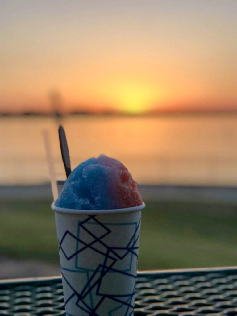 A patterned cup of blue and red shaved ice with a straw is silhouetted against a blurred sunset over the water.