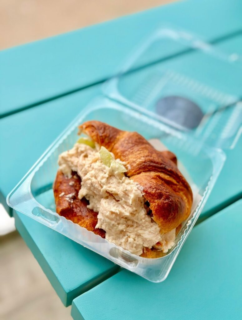 A chicken salad sandwich served on a flaky, golden-brown croissant inside a clear plastic container resting on a bright turquoise wooden table.