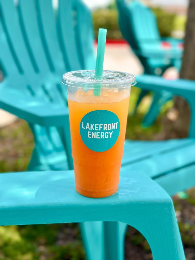 A clear plastic cup filled with an orange energy drink sits on a bright turquoise table, with turquoise Adirondack chairs in the blurred background. A turquoise straw sticks out of the lid, and a round sticker on the cup reads "LAKEFRONT ENERGY".