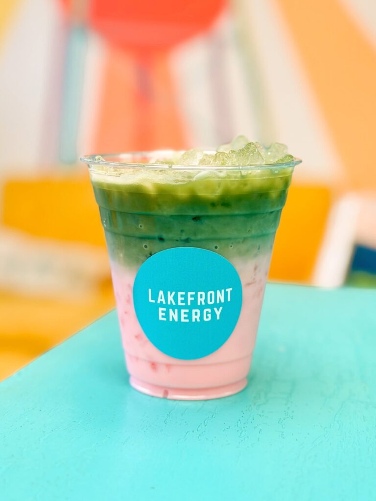An iced drink in a clear plastic cup sits on a bright blue surface, featuring layered colors of pink at the bottom, white in the middle, and vibrant green at the top. A circular turquoise sticker on the cup features the text "LAKEFRONT ENERGY" in white. The background is softly blurred with warm yellow, orange, and white tones.