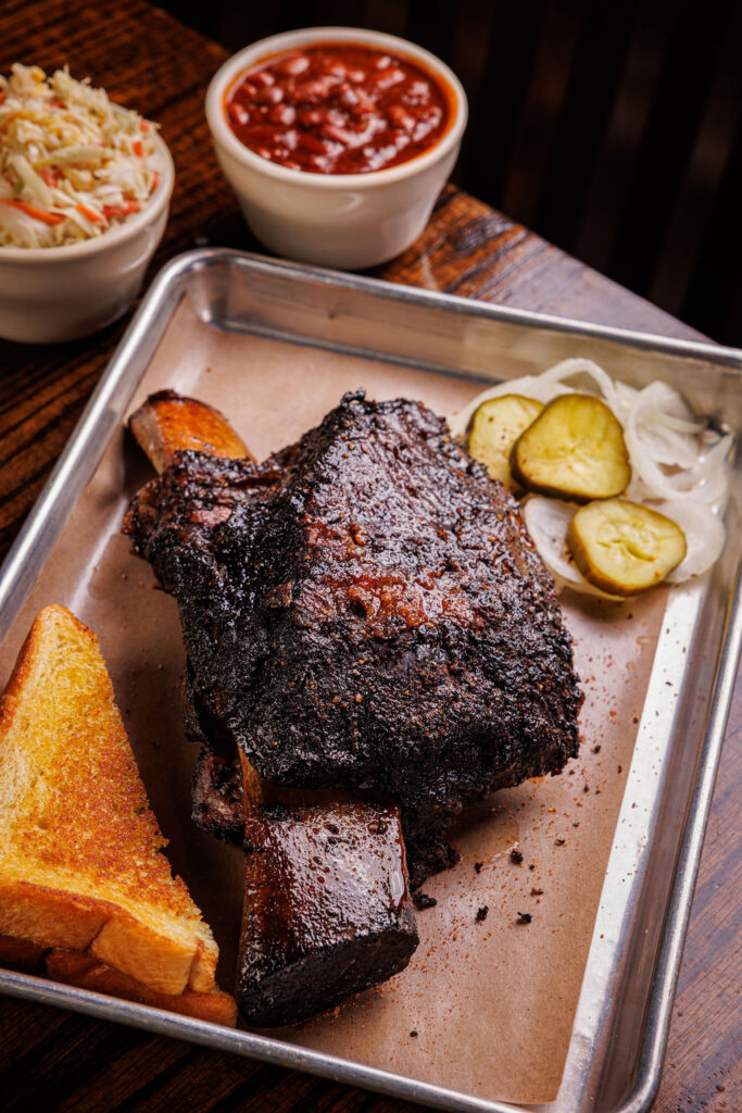 Barbeque Rib on a platter with pickles and toast, with a side of coleslaw and beans.