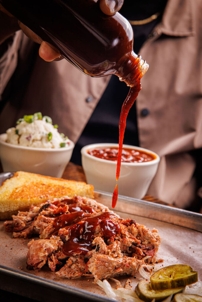 A photo of someone pouring barbeque sauce over a plate of brisket, toast and pickles. With a side of potato salad and beans in the background.