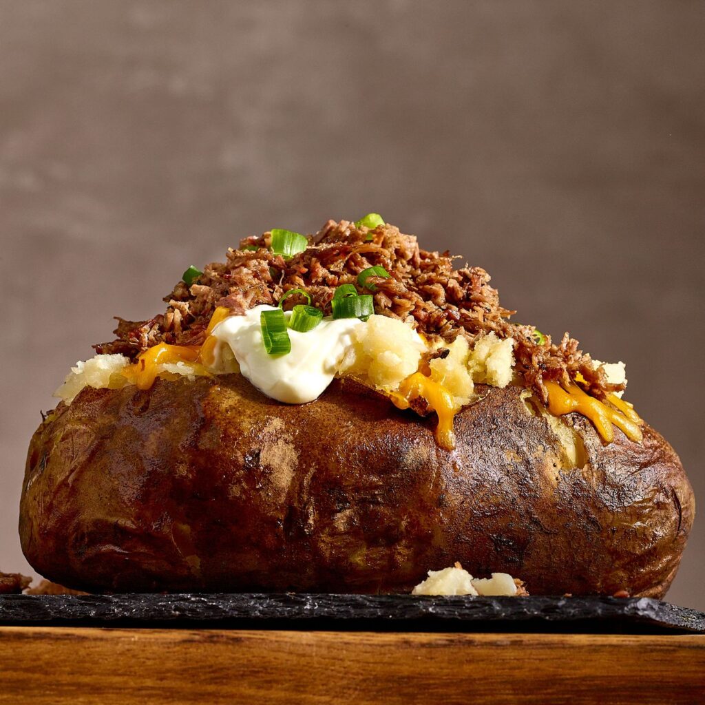 Baked potato topped with cheese, brisket, sour cream, and chives, placed on top of a cutting board, against a grey background.