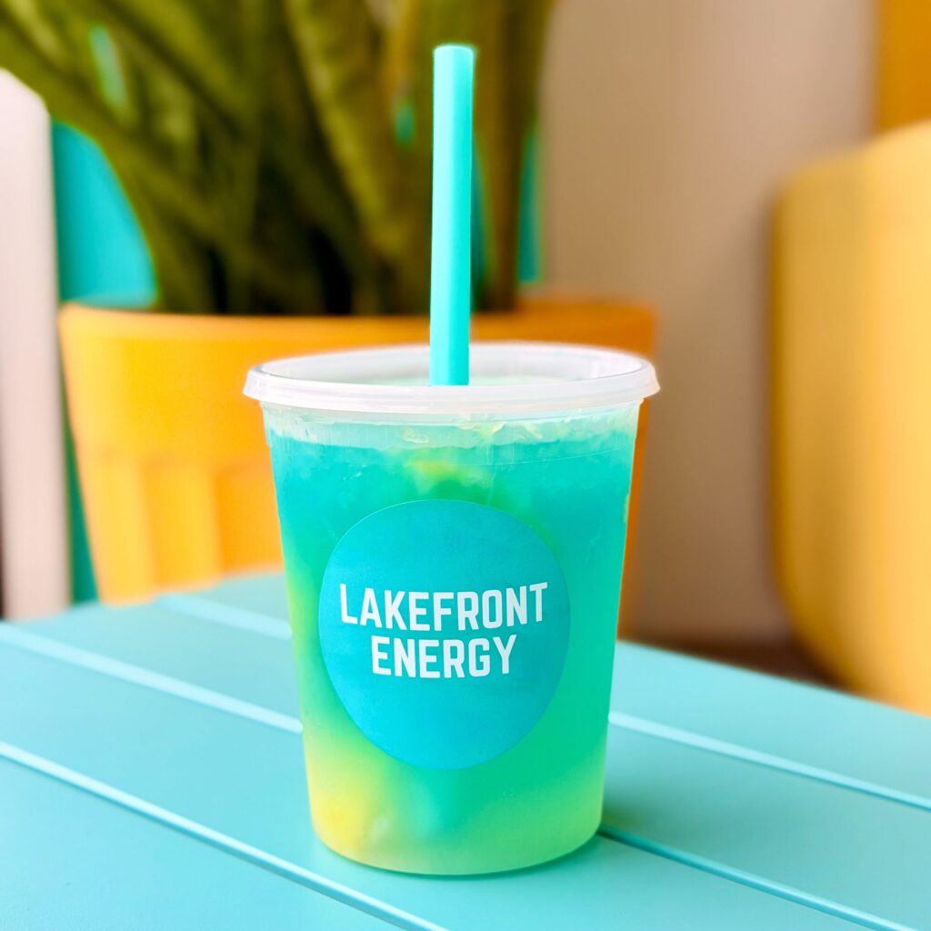 A clear plastic cup of turquoise-blue and yellow energy drink with a matching turquoise straw sits on a blue table. A round sticker on the cup reads 'LAKEFRONT ENERGY' in white capital letters. In the soft-focus background, a yellow planter holds a green plant against a teal wall.