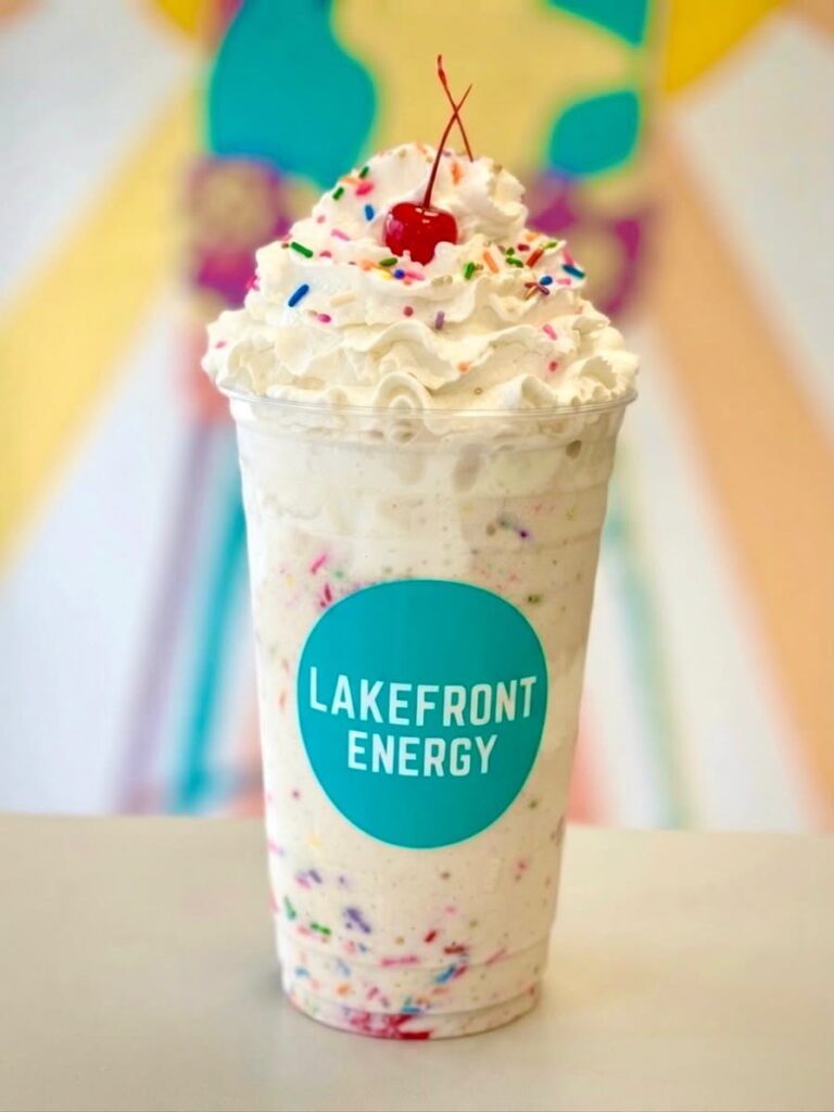 A tall clear plastic cup filled with a funfetti milkshake, topped with a generous mound of whipped cream, colorful sprinkles, and a single bright red cherry with its stem, set against a blurred colorful background and bearing a turquoise circular logo that reads "Lakefront Energy".