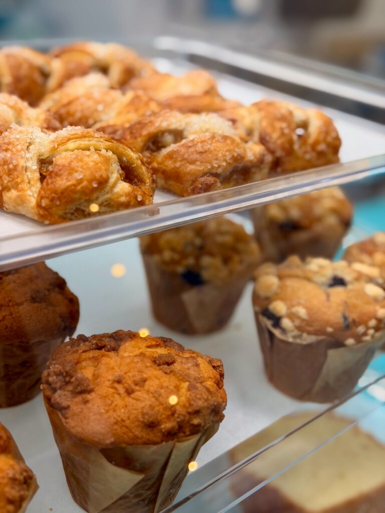 An eye-level close-up shows a bakery display case filled with rows of golden pastries and muffins. The top shelf holds glazed pastries dusted with sugar, while the bottom shelf displays several muffins with crumble toppings in paper wrappers, all shot with a shallow depth of field.