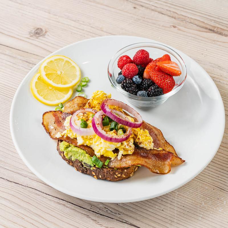 Toast topped with avocado, scrambled eggs, bacon, red onion, and green onion sits beside lemon slices and a bowl of mixed berries.