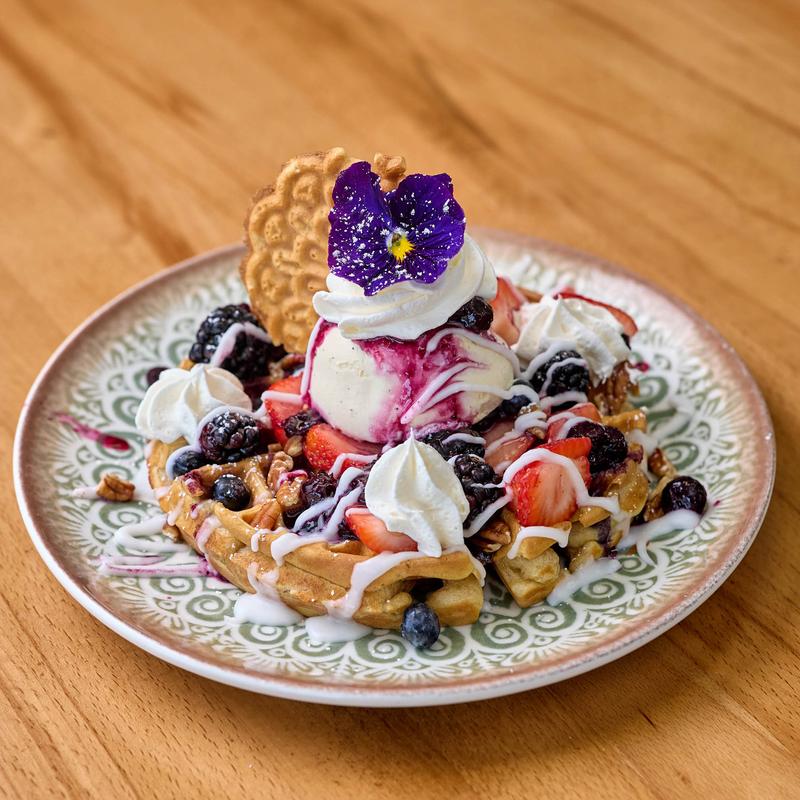 A plate of waffles topped with ice cream, whipped cream, berries, nuts, white drizzle, a wafer, and a purple flower.