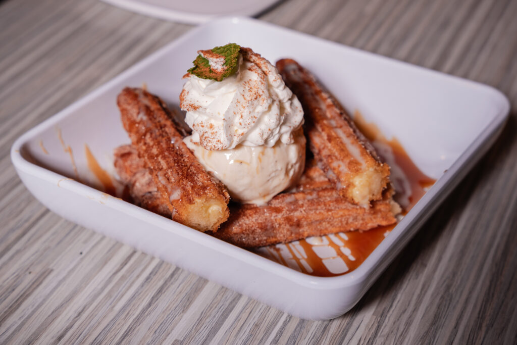 A close-up shot of a white square bowl filled with crispy, golden churros dusted in cinnamon sugar, served with a scoop of vanilla ice cream, whipped cream, and a drizzle of caramel syrup on a textured wooden table.