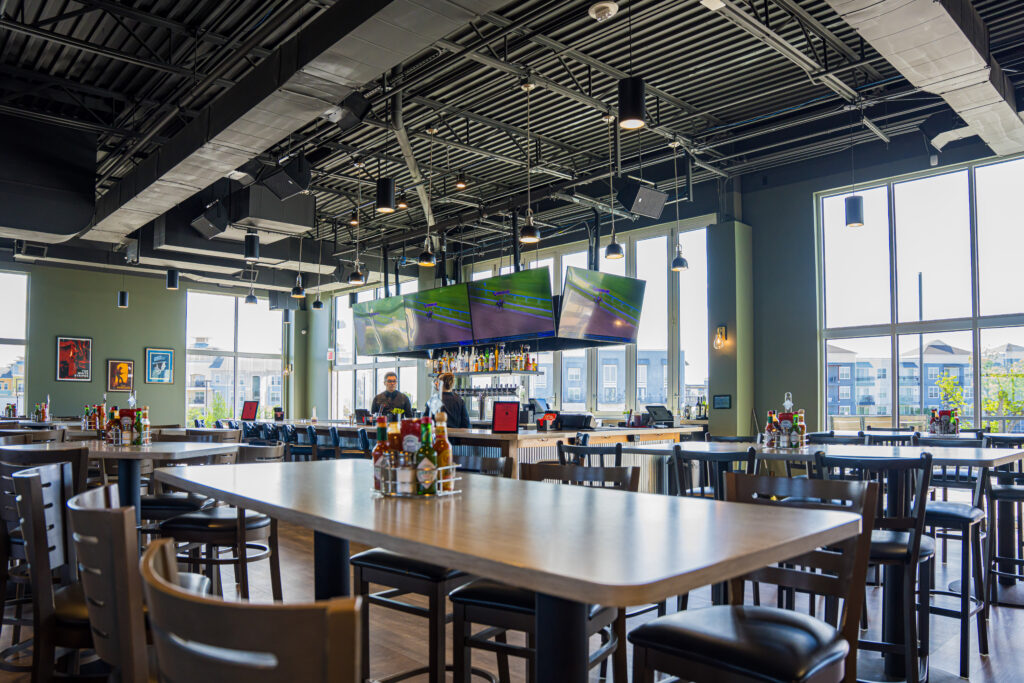 An interior shot of a modern restaurant featuring high ceilings with exposed ductwork, large windows providing natural light, and several flat-screen TVs mounted above the bar. Tables and chairs fill the foreground while staff members work behind the bar counter.