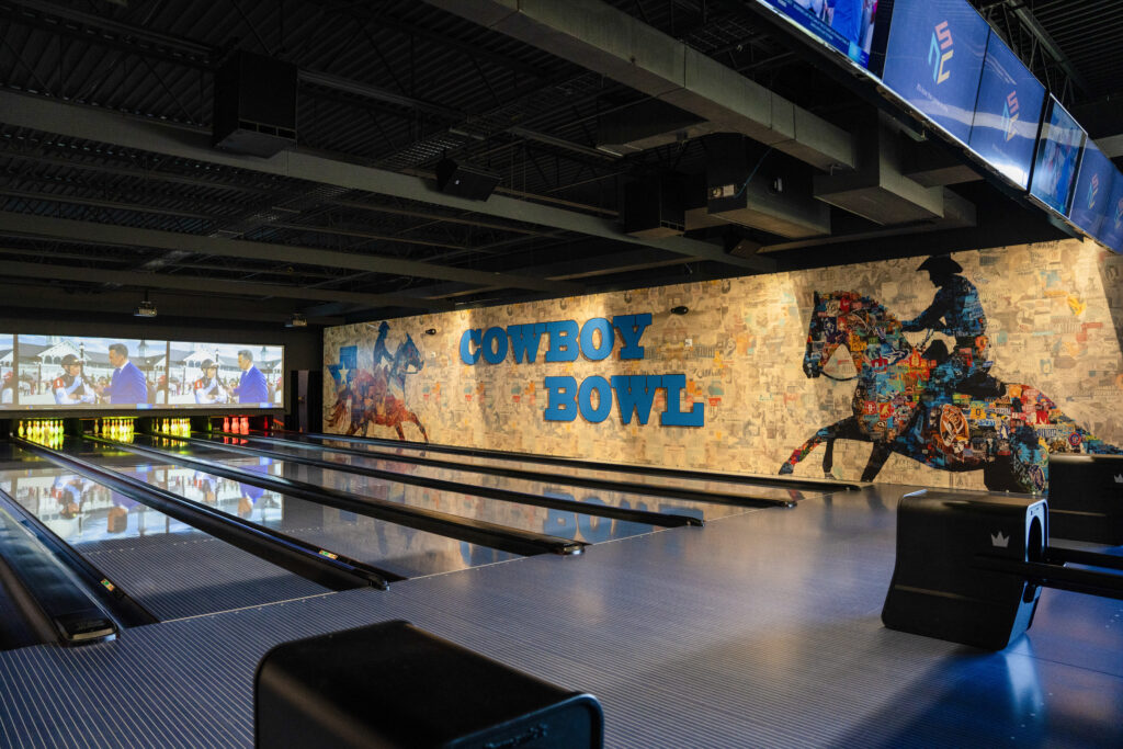 An indoor bowling alley with multiple lanes stretching into the distance. The back wall features a large collage mural of a cowboy riding a horse alongside the blue text reading 'COWBOY BOWL'.