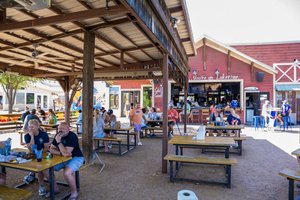 A sports bar with wooden tables and metal chairs, patrons at the counter, televisions showing sports, and Bud Light signs overhead.